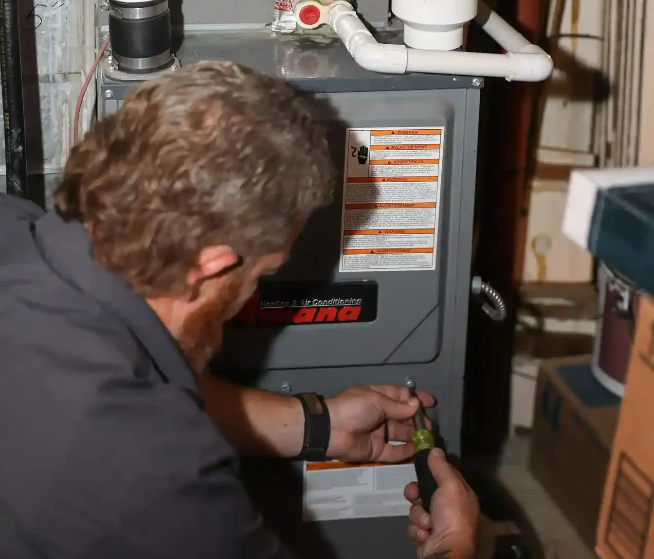Technician performs routine maintenance on an Amana furnace, inspecting components using a screwdriver in a utility room.