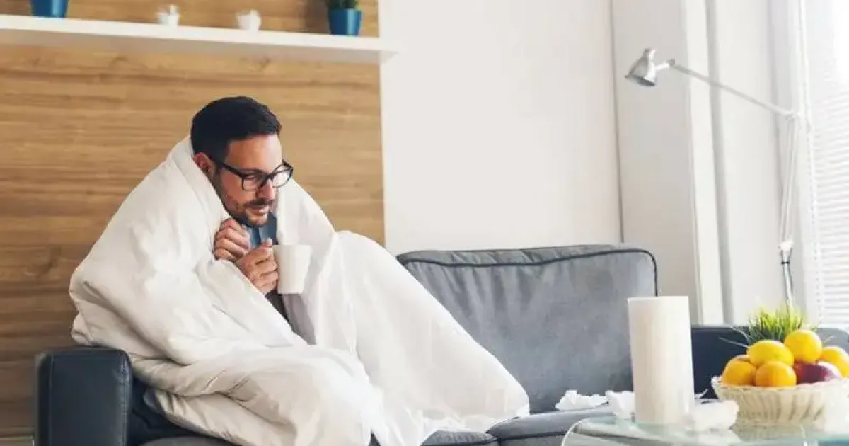 Cold man on a couch with a mug and blanket in a home with a heat pump blowing cold air.