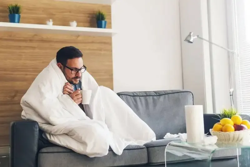 Cold man on a couch with a mug and blanket in a home with a heat pump blowing cold air.