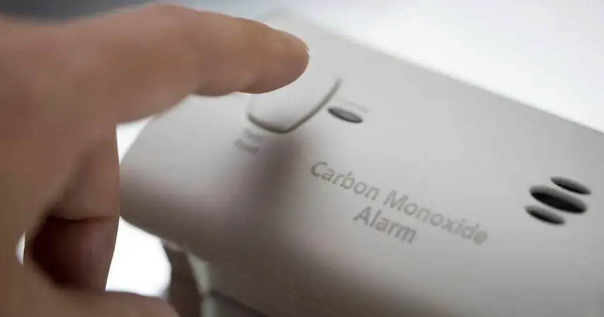 Hand pressing alarm button on a white carbon monoxide alarm.