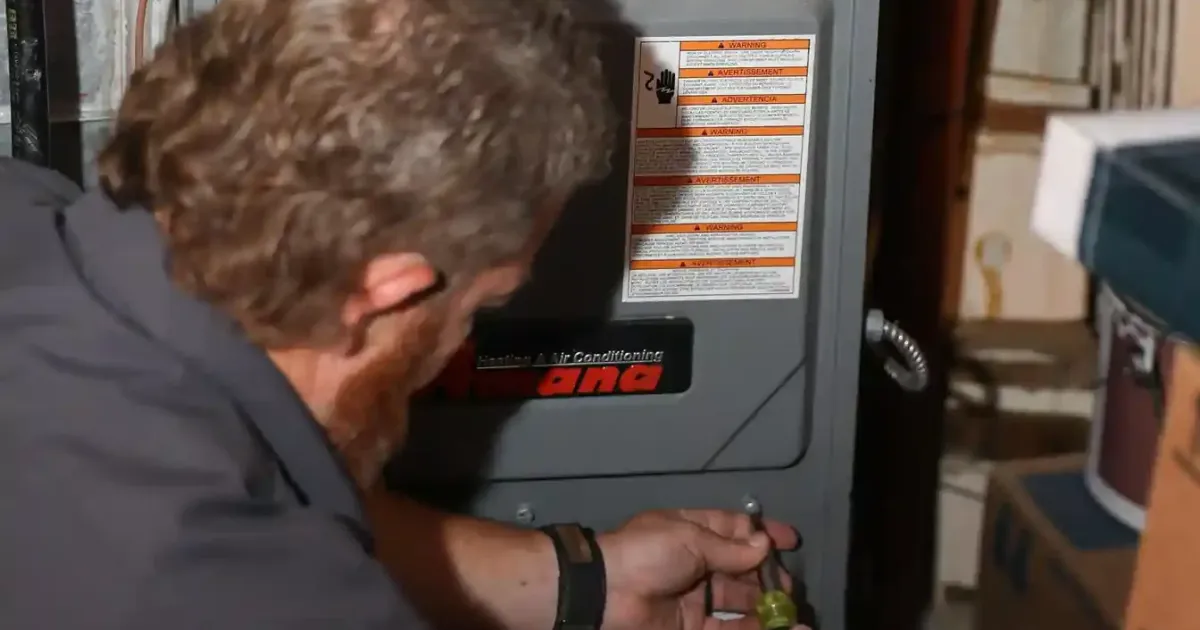 Technician performs routine maintenance on an Amana furnace, inspecting components using a screwdriver in a utility room.