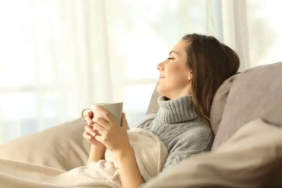 Woman relaxing under a blanket, holding a mug while reclining on a couch.