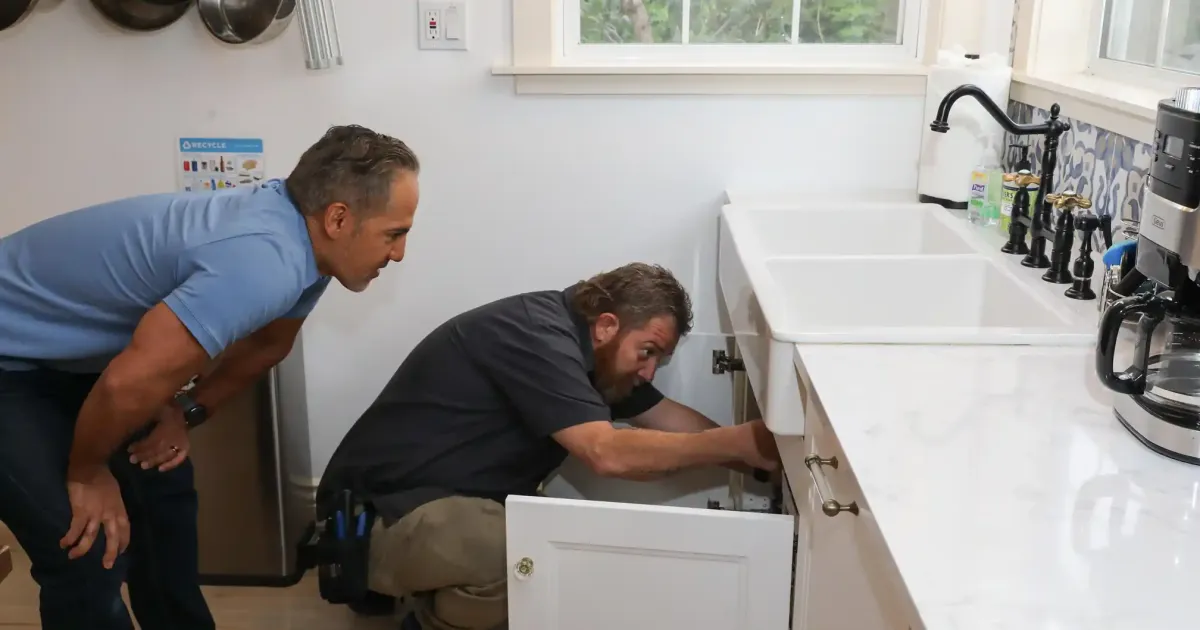 Plumber works under a kitchen sink in a modern home, performing focused repair or maintenance activities.