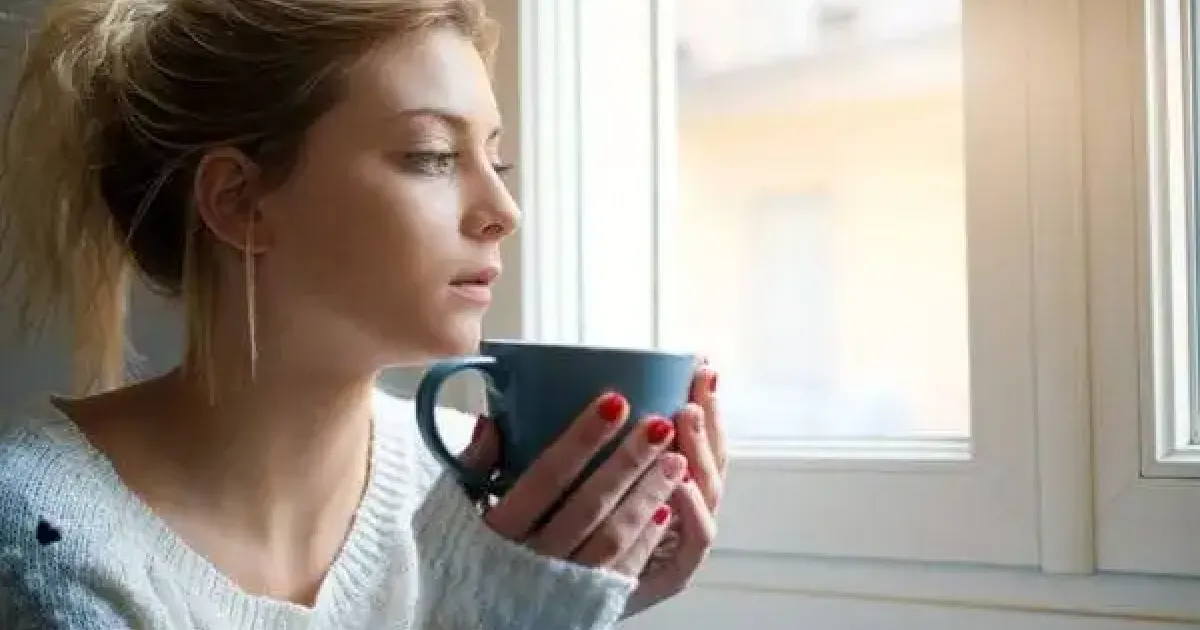 A woman sits with a cup by a window.