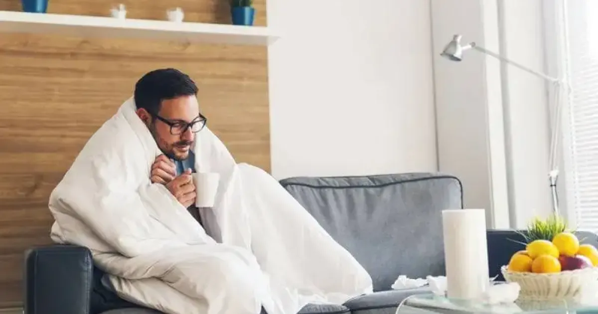 Cold man on a couch with a mug and blanket in a home with a heat pump blowing cold air.