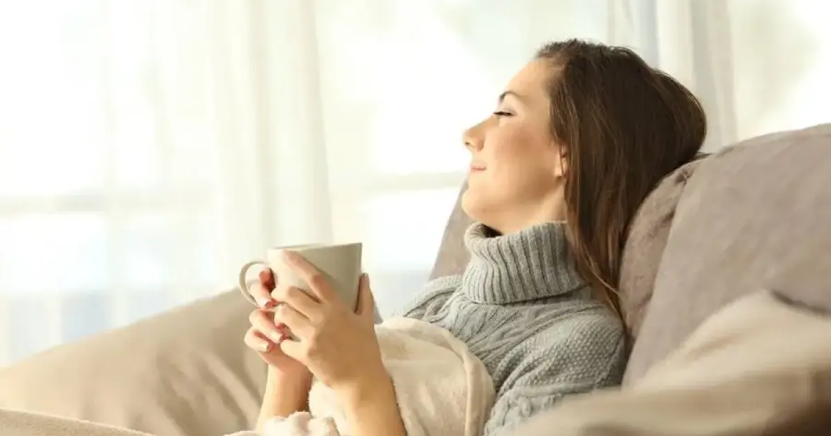 Woman relaxing under a blanket, holding a mug while reclining on a couch.